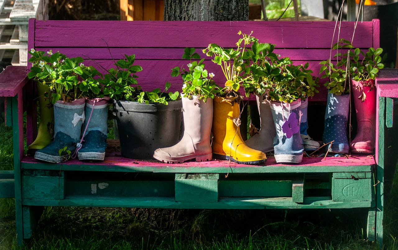 hero-img-02 Vibrant rain boots repurposed as planters with lush greenery on a painted garden bench.